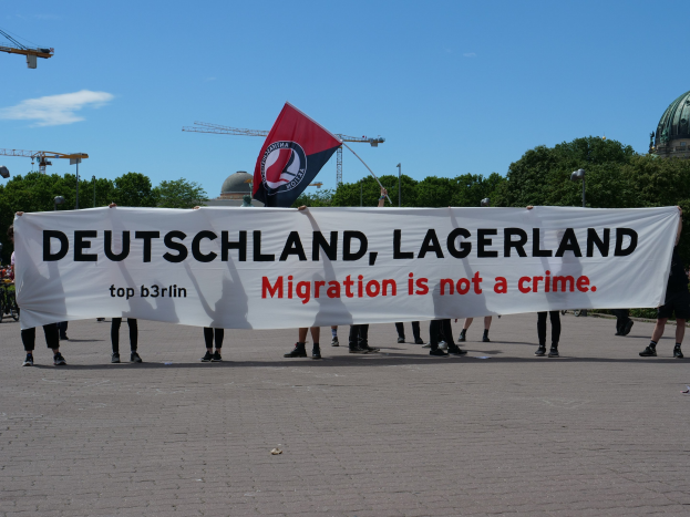 Gruppe von Menschen mit einer Flagge und einem Transparent mit der Aufschrift "Deutschland, Lagerland Migration ist kein Verbrechen" sowie einer Straßeninfrastruktur, Bäumen, einem Gebäude und einem bewölktem Himmel im Hintergrund.