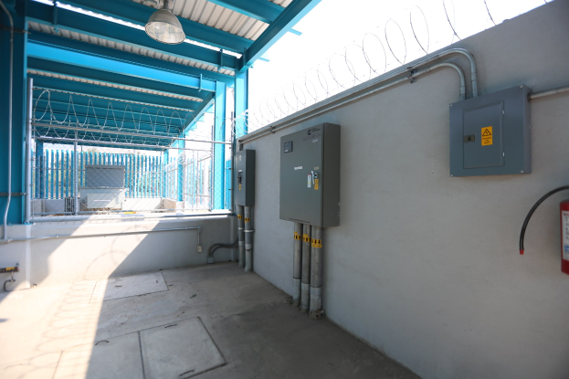 Ein Raum mit einem Feuerlöscher auf der rechten Seite, elektrischen Schaltern und Rohren an der Wand, einer Deckenlampe und einem Zaun im Hintergrund, was auf eine elektrische Unterstation hindeutet.