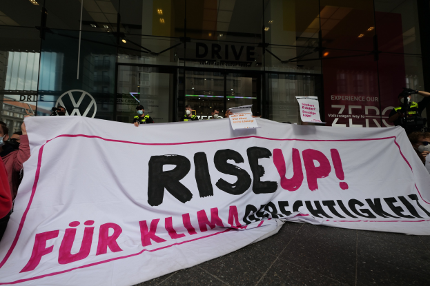 Eine Gruppe von Menschen mit Masken hält ein Transparent mit der Aufschrift "Aufstehen" und Schilder vor einem Glasgebäude, um gegen eine Entscheidung der deutschen Regierung bezüglich Frauenrechten zu protestieren.