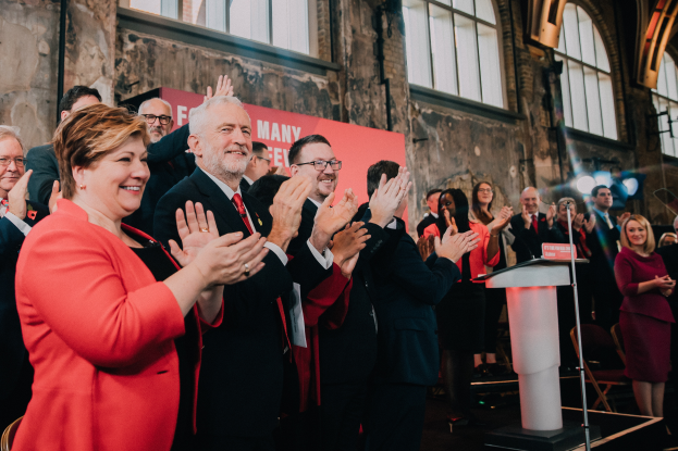 Eine Gruppe von Menschen, wahrscheinlich FDP-Mitglieder, steht vor einer Menge, klatscht feierlich, mit einem Podium, Mikrofon und Texttafel rechts und Stühlen, Banner, Wand, Fenstern und Lichtern im Hintergrund.