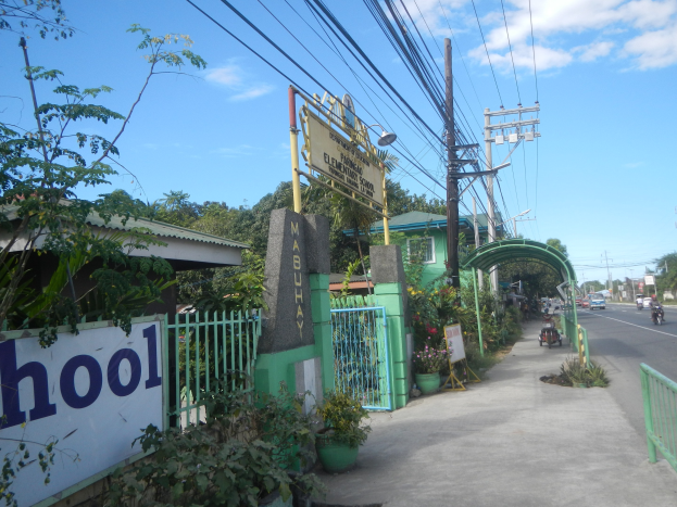 Schuleingang mit einem "Schule"-Schild am Straßenrand, mit einem Tor, einem Bogen, einer Namensplatte, Fahrzeugen, Bäumen, Pflanzen, Strommasten mit Drähten und einem bewölkten Himmel.