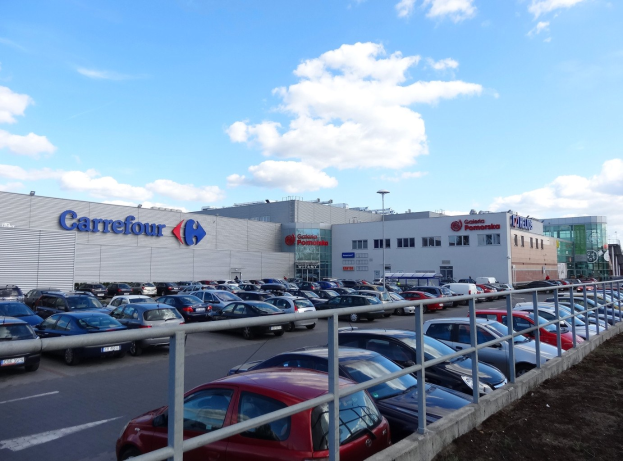 Großes Autohaus mit zahlreichen geparkten Fahrzeugen, umgeben von Gebäuden, Straßeninfrastruktur und Fahrzeugen auf der Straße unter einem bewölkten Himmel.