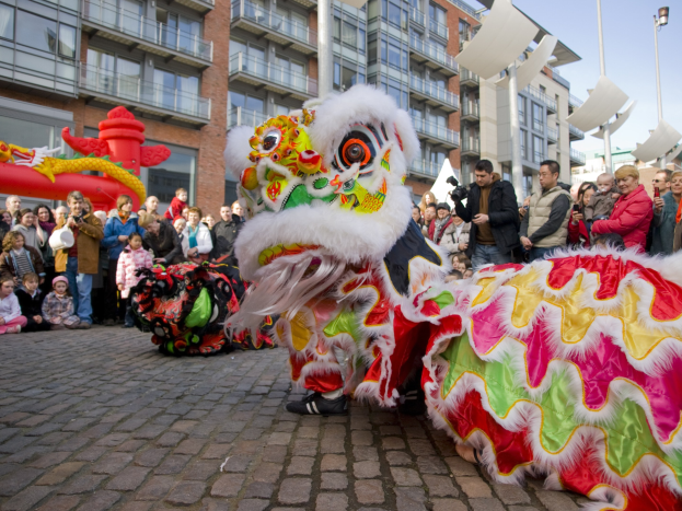 Ein farbenfrohes chinesisches Neujahrsfest in Amsterdam mit einem Löwen tanzen im Vordergrund und einer Menge Menschen, einige mit Kameras, sowie Gebäuden, Laternenmasten und einem klaren blauen Himmel im Hintergrund.
