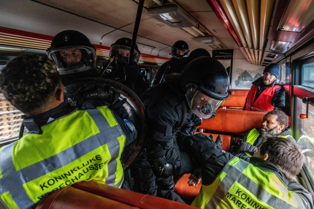 Eine Gruppe von Polizisten in Einsatzausrüstung auf einem Bus, einige tragen Helme und Jacken, mit einer Person, die in der Mitte sitzt, und einem Plakat an der Wand im Hintergrund.