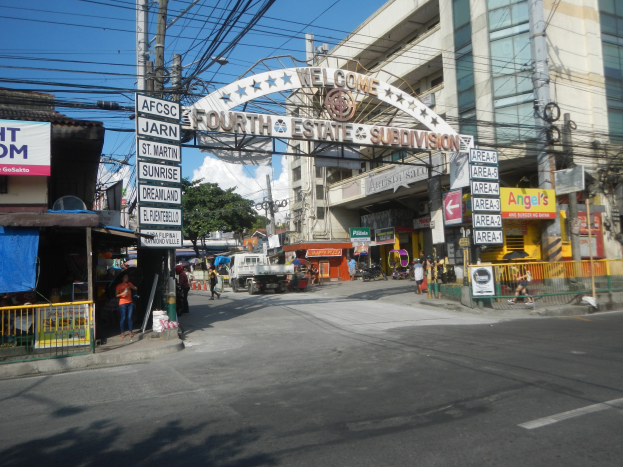 Eine belebte Stadtstraße mit Fahrzeugen, Fußgängern, Gebäuden, Bäumen und einem Schild mit der Aufschrift "Vierter Stand" in der Mitte, unter einem bewölkten Himmel.