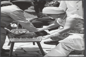 Ein Schwarz-Weiß-Foto eines Mannes und einer Frau, die auf einem Tisch Backgammon spielen, umgeben von einer Gruppe von Menschen, Pflanzen und Gras.