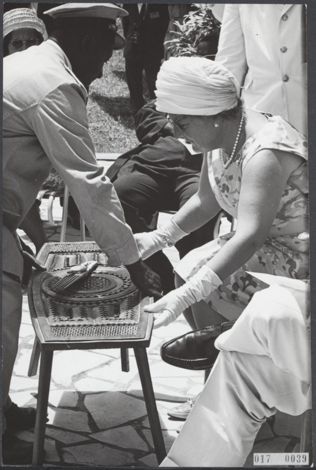 Ein Schwarz-Weiß-Foto eines Mannes und einer Frau, die auf einem Tisch Backgammon spielen, umgeben von einer Gruppe von Menschen, Pflanzen und Gras.