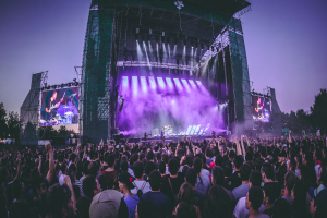 Eine große Menschenmenge versammelte sich auf einem Musikfestival, mit einer Bühne in der Mitte, auf der Musiker Instrumente spielten, beleuchtet von Scheinwerfern und umgeben von Bildschirmen, und einem Gebäude, Bäumen und einem klaren blauen Himmel im Hintergrund.