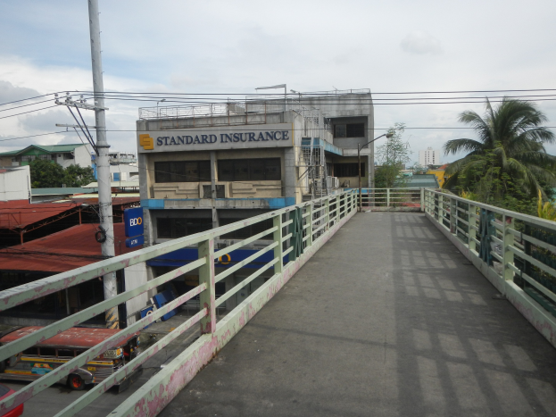 Eine Brücke mit Geländern, Fahrzeugen, Gebäuden, Bäumen, Strommasten mit Leitungen und einem bewölktem Himmel im Hintergrund, mit einem Gebäude an der Seite mit einer "Standard Insurance"-Aufschrift.