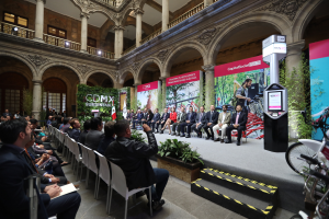 Eine Gruppe von Menschen sitzt in Stühlen und schaut auf einen Mann auf einer Bühne, der ein Mikrofon hält, mit Fahrrädern rechts geparkt und verschiedenen Elementen wie Plakaten, Pflanzen, Säulen, Geländern, Lampen, Fenstern und einer Wand im Hintergrund.