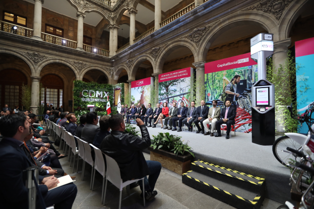 Eine Gruppe von Menschen sitzt in Stühlen und schaut auf einen Mann auf einer Bühne, der ein Mikrofon hält, mit Fahrrädern rechts geparkt und verschiedenen Elementen wie Plakaten, Pflanzen, Säulen, Geländern, Lampen, Fenstern und einer Wand im Hintergrund.