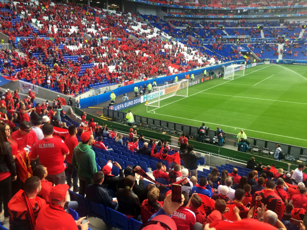 Eine große Menschenmenge in einem Stadion schaut einem Fußballspiel zu, mit Menschen, die sitzen und stehen, Bannern, einem Torpfosten und Deckenlampen.