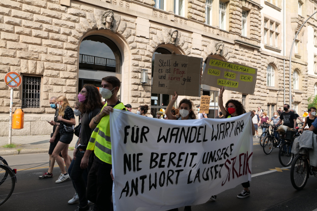 Eine Gruppe von Menschen, die an einer Protestdemo durch die Straße marschieren, einige halten Schilder und andere fahren Fahrräder, mit einem Gebäude im Hintergrund, das Fenster, Bögen, Säulen und Skulpturen aufweist.