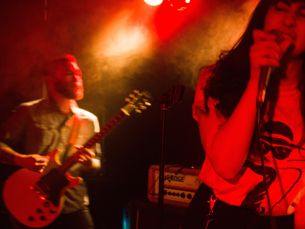 Eine Frau singt in ein Mikrofon auf der Bühne, wöhrend ein Mann eine Akustikgitarre im Hintergrund spielt, mit Deckenlampen, die die Bühne beleuchten.