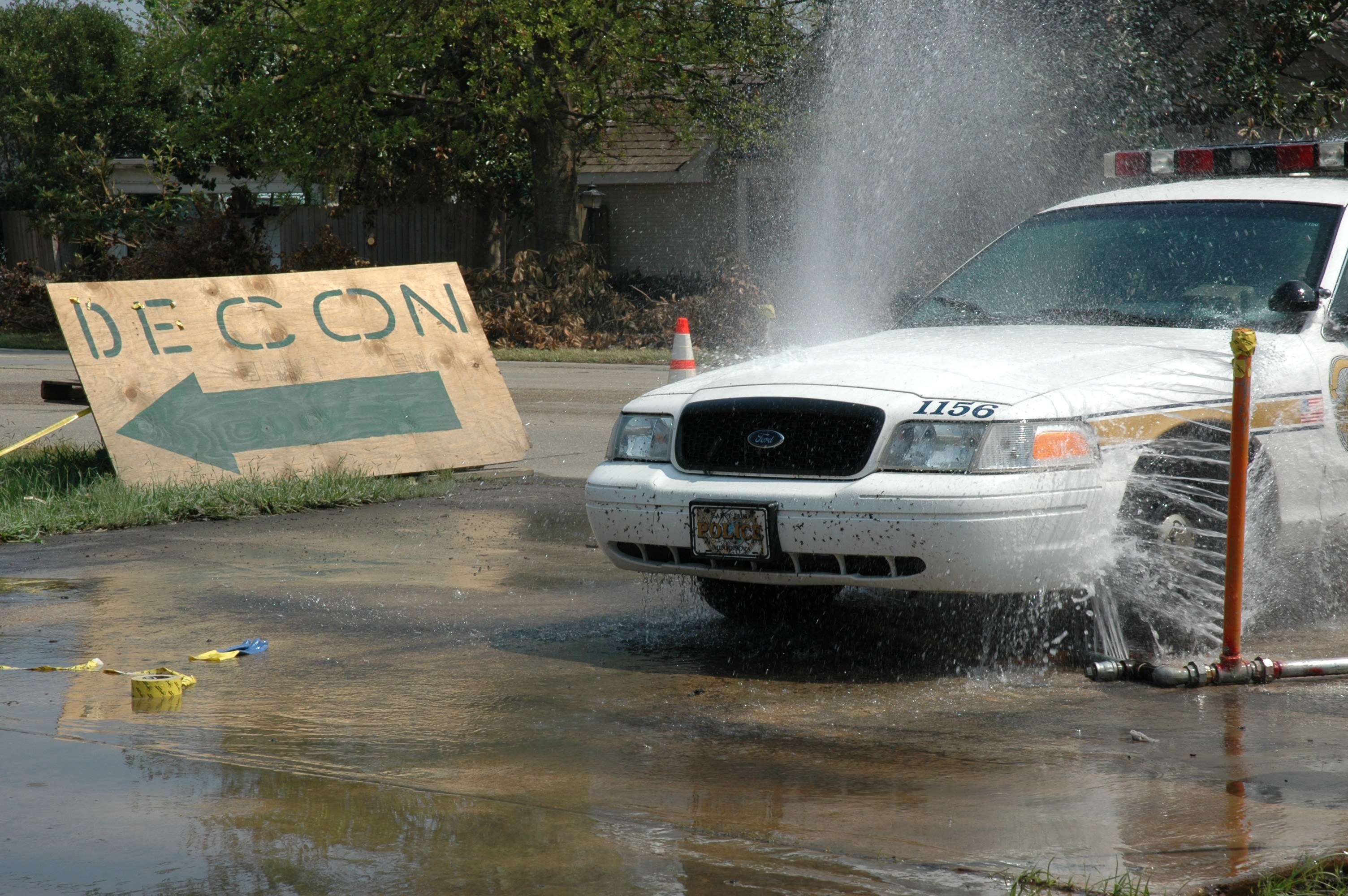 Ein Polizeiwagen fährt durch eine Pfütze mit verstreuten Gegenständen, darunter eine Tafel mit Text und ein Verkehrskegel, im Hintergrund Bäume und Gebäude unter einem klaren blauen Himmel.