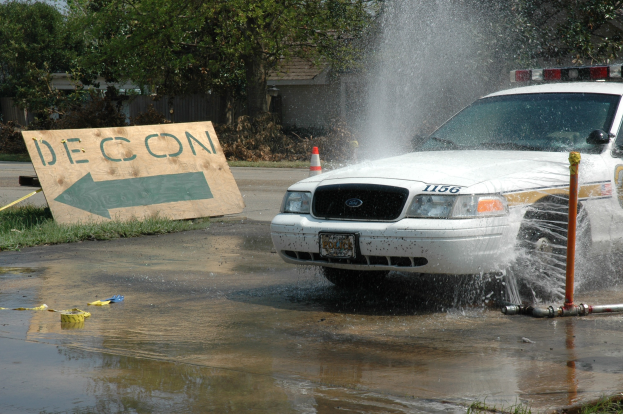 Ein Polizeiwagen fährt durch eine Pfütze mit verstreuten Gegenständen, darunter eine Tafel mit Text und ein Verkehrskegel, im Hintergrund Bäume und Gebäude unter einem klaren blauen Himmel.