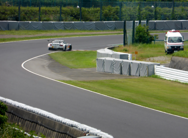 Ein Rennauto fährt eine Rennstrecke entlang mit einem Polizeiwagen im Hintergrund, umgeben von Gras, Pflanzen, Bäumen und Zaun, mit einem Fahrzeug auf der rechten Seite und Text unten.