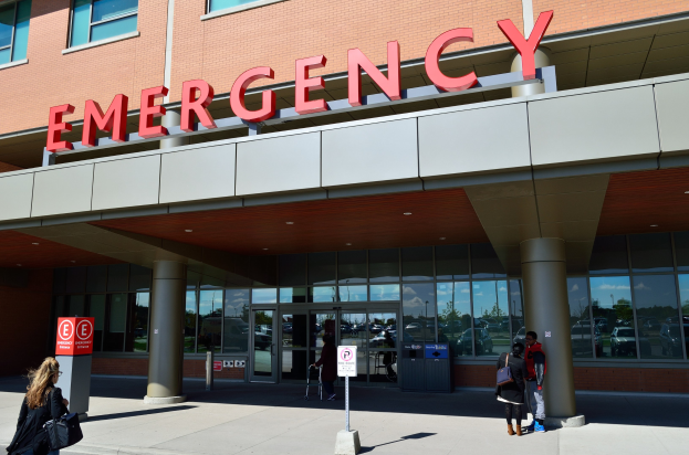 Gruppe von Menschen, die auf ein Notaufnahmegebäude mit Glaswänden und -säulen zusteuert, mit einem Namensschild, Hinweisschildern und einem Müllimer.