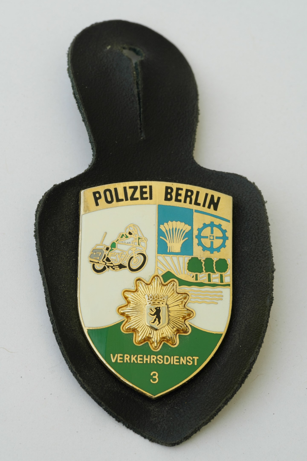 Ein schildförmiges Polizeibadged mit 'Polizei Berlin' in schwarzer Schrift auf einem blauen Hintergrund, das ein weißes Stern in der Mitte und einen weißen Rand hat.