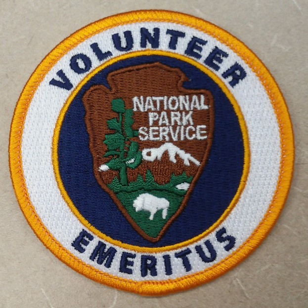 Runder Aufnäher mit "Volunteer National Park Service Emeritus" in fetter schwarzer Schrift auf einem weißen Hintergrund, umrandet von einem blauen Rand und einem gelben Kreis.