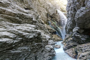 Kleiner Wasserfall, der über zerklüftete Felsen in einem steinigen Tal hinabstürzt, umgeben von grünen Hügeln, unter strahlendem Sonnenlicht mit klaren Wasser.