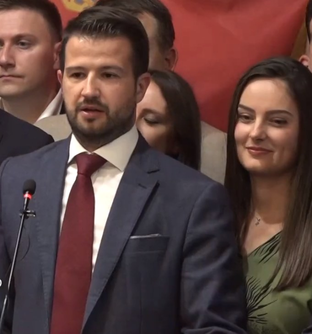 Ein Mann im Anzug steht an einem Rednerpult und spricht in ein Mikrofon, mit einem Banner im Hintergrund und einer Menge vorne, wahrscheinlich zu einem sozialen Medienverbot der Regierung.