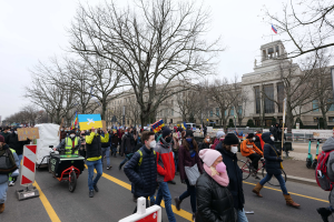 Eine große Gruppe von Menschen nimmt an einer Protestdemo auf einer Straße in Washington, D.C. teil, einige halten Schilder und Transparente, andere fahren Fahrräder, Schilder mit Stangen, Bäume und ein klarer blauer Himmel im Hintergrund.
