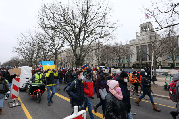 Eine große Gruppe von Menschen nimmt an einer Protestdemo auf einer Straße in Washington, D.C. teil, einige halten Schilder und Transparente, andere fahren Fahrräder, Schilder mit Stangen, Bäume und ein klarer blauer Himmel im Hintergrund.
