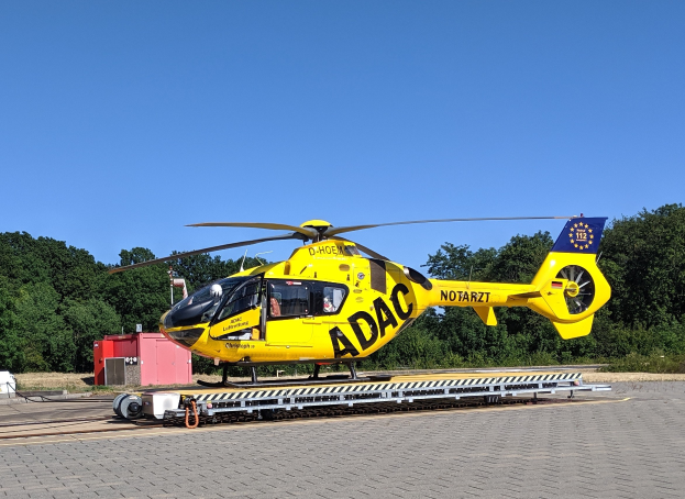 Gelber Hubschrauber auf einem Flughafenvorfeld mit umliegenden Bäumen und einem klaren blauen Himmel, wahrscheinlich ein Luftrettungshubschrauber in Deutschland.
