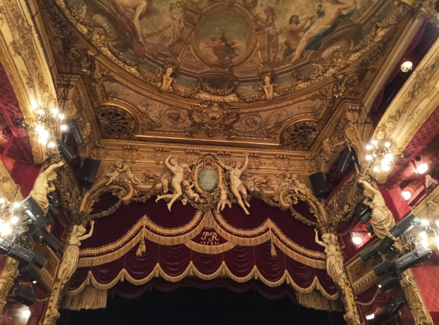 Theaterinnenraum mit roter Gardine, Kronleuchtern, Deckenplastiken, Lichtern und Gemälde, mit einer Bühne und einer kleinen Gruppe von Menschen unten.