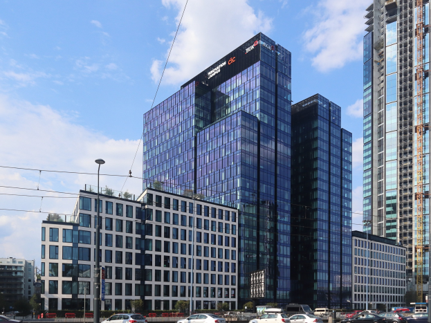 Headquarters of Deutsche Bank in Frankfurt, Germany, a large multi-windowed building surrounded by urban infrastructure including street poles, vehicles, and greenery under a cloudy sky.