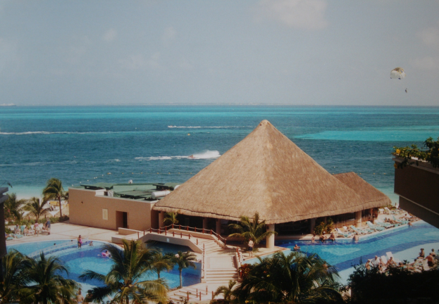 Resort mit einem gro├čen Schwimmbad umgeben von Palmen, Hütten und Menschen, überblickend auf ein Meer unter einem bew├Âlktem Himmel mit einem Fallschirm in der Luft.