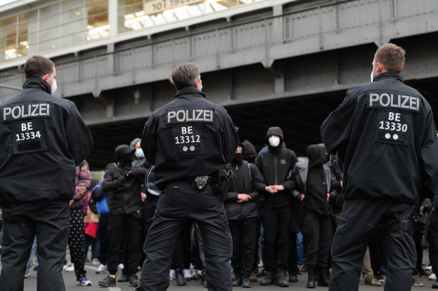 Eine Gruppe von Polizisten in schwarzen Uniformen und Masken steht vor einer Menge, mit einer Stadtbrücke und einem Gebäude im Hintergrund, während einer Demonstration.