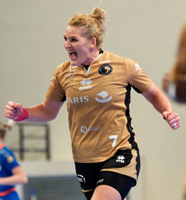 Eine Frau in brauner Bluse und schwarzen Shorts rennt auf einem Futsalplatz, feiernd mit triumphierendem Gesichtsausdruck, während zwei andere Frauen im Hintergrund zu sehen sind.
