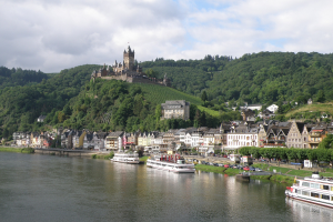 Ein malerischer Blick auf den Rhein in Deutschland mit einer Burg auf einem Hügel, Booten auf dem Fluss, Fahrzeugen auf einer nahen Straße und einem bewölkten Himmel.