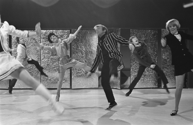 Eine Gruppe von Menschen, die auf einer Bühne in Schwarz-Weiß tanzen, tragen Schuhe und haben eine Wand im Hintergrund.