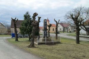 Ein kleines Dorf mit einem Friedhof in der Mitte, umgeben von Bäumen, Gras, einem Zaun, einer Straße, Fahrzeugen, Mülltonnen und Gebäuden mit Fenstern unter einem bewölkten Himmel, mit einem Gedenkstein mit eingravierter Schrift auf dem Boden.