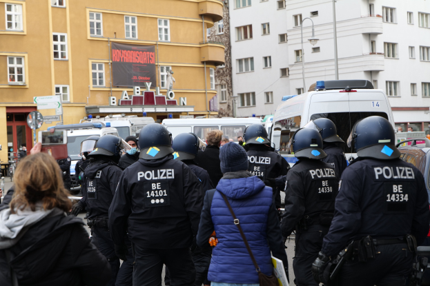 Polizeibeamte in Uniform vor einer Menge von Helmträgern während einer Demonstration in Berlin, mit Fahrzeugen, Gebäuden und einem Banner im Hintergrund.