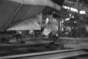 Ein Schwarz-Weiß-Foto eines Boots in der Fertigung in einer Fabrik, umgeben von metallenen Gegenständen, Pfählen und Lichtern.