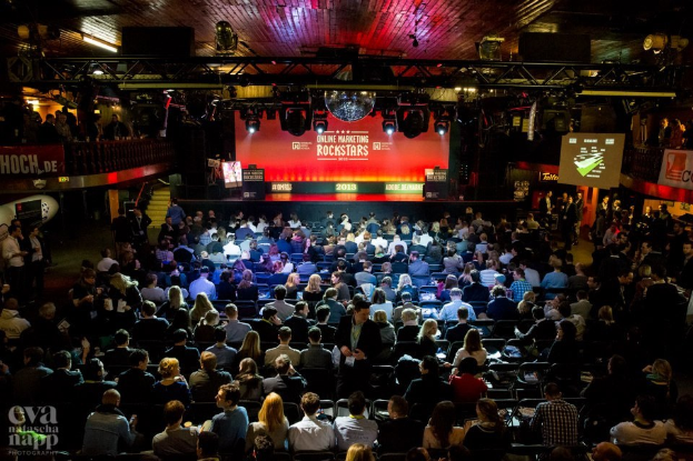 Eine große Menschenmenge, die auf Stühlen sitzt und einer hell erleuchteten Bühne mit Bildschirmen und Bannern gegenübersitzt, was eine Vorfreude auf ein Online-Marketing-Event vermuten lässt.