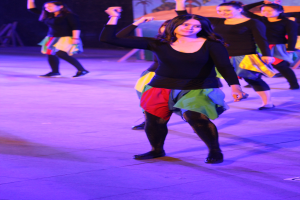 Eine Gruppe junger Frauen tanzt auf einer Bühne mit einem Wandgemälde im Hintergrund, alle tragen Schuhe.