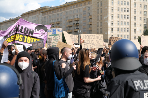 Eine Gruppe maskierter Demonstranten mit Schildern vor einem Gebäude, mit zweihelmigen Polizisten rechts daneben, unter einem bewölkten Himmel mit Bäumen im Hintergrund.