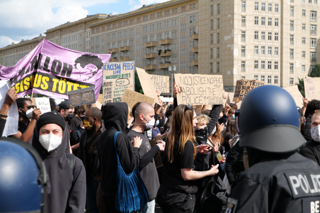 Eine Gruppe maskierter Demonstranten mit Schildern vor einem Gebäude, mit zweihelmigen Polizisten rechts daneben, unter einem bewölkten Himmel mit Bäumen im Hintergrund.