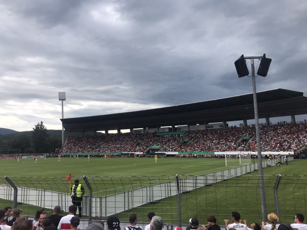 Große Zuschauermenge bei einem Fußballspiel in einem bewölkten Stadion mit beleuchtetem Feld, umgeben von einem Zaun, Bäumen, Hügeln und Laternenmasten.