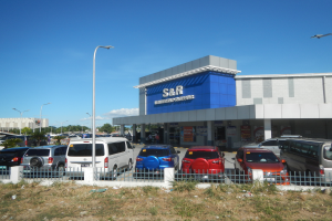 Ein Autohaus mit parkenden Fahrzeugen vorne, umgeben von einem Metallzaun, Gras, Pflanzen, Bäumen, Straßenlaternen und einem bewölkten Himmel.