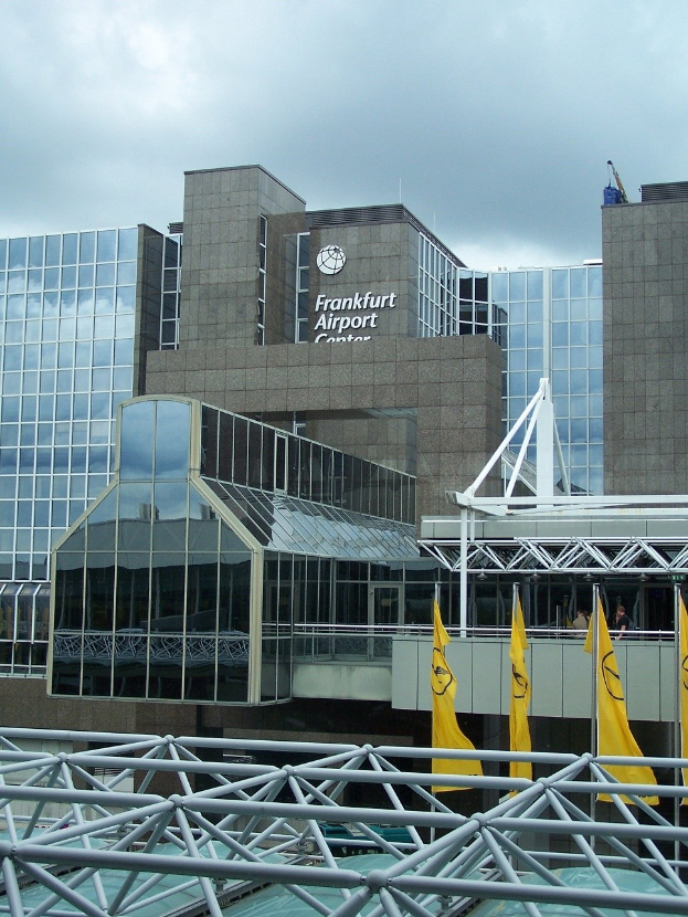 Frankfurt Airport in Frankfurt, Germany, featuring a large glass building with text, surrounded by yellow flags and iron rods under a cloudy sky.
