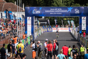 Eine Gruppe von Radfahrern fährt eine Straße entlang, die von Zuschauern gesäumt ist, unter einem Bogen, auf dem "2019 UEC MTB Europameisterschaften" steht, mit Bäumen, Gebäuden und einem klaren blauen Himmel im Hintergrund.