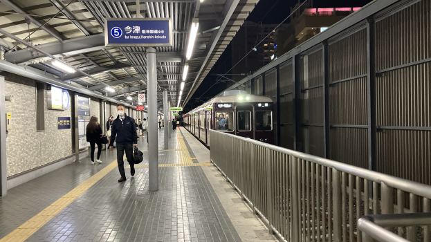 Ein Zug fährt auf einem Bahnsteig ein, mit Menschen in der Nähe, einem Geländer auf der rechten Seite, Gebäuden im Hintergrund, Bahnsteiglücken und einer Abfahrtsanzeige.