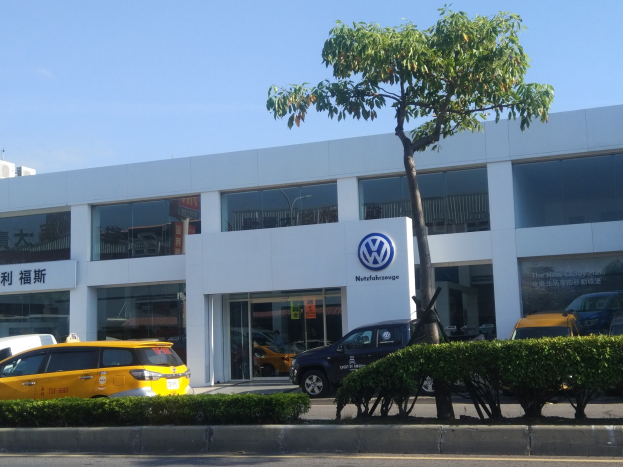 Ein VW-Autohaus mit parkenden Autos vorne, umgeben von Pflanzen und einem Baum, mit Glaswänden und -fenstern vor einem Himmelhintergrund.