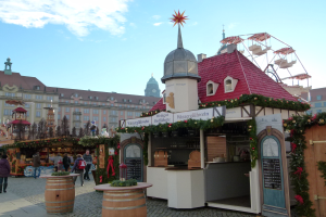 Ein geschäftiger Weihnachtsmarkt in Nürnberg, Deutschland mit Menschen um geschmückte Stände, festliche Lichter, ein Riesenrad, Gebäude und eine Tafel mit Text auf der rechten Seite.